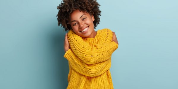 Lovely happy African American woman daydreams, embraces herself, recalls romantic date, feels comfort, keeps hands across body sensually, has broad smile, wears yellow sweater, poses indoor.